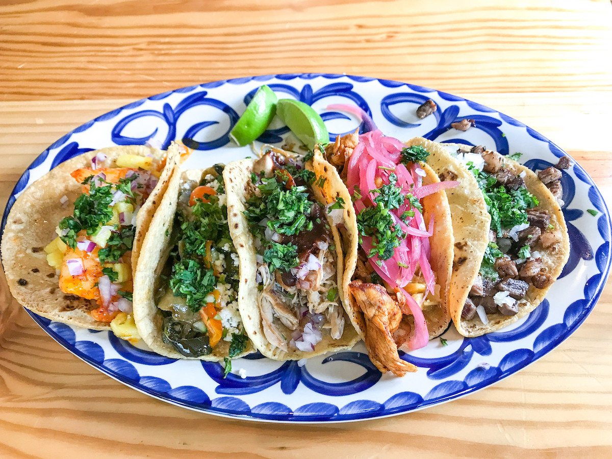 A wooden table with a blue and white painted platter on top of it with five tacos and lime wedges