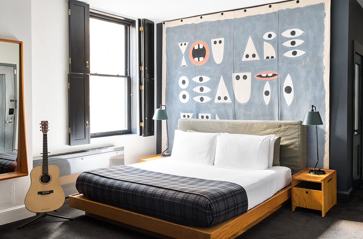 queen sized bed in a sunlit room with a guitar nearby and a large art piece on the wall.