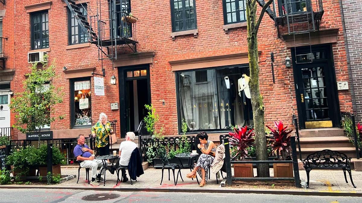 tall brick building with large windows, iron tables and chairs, and plants growing along the sidewalk.