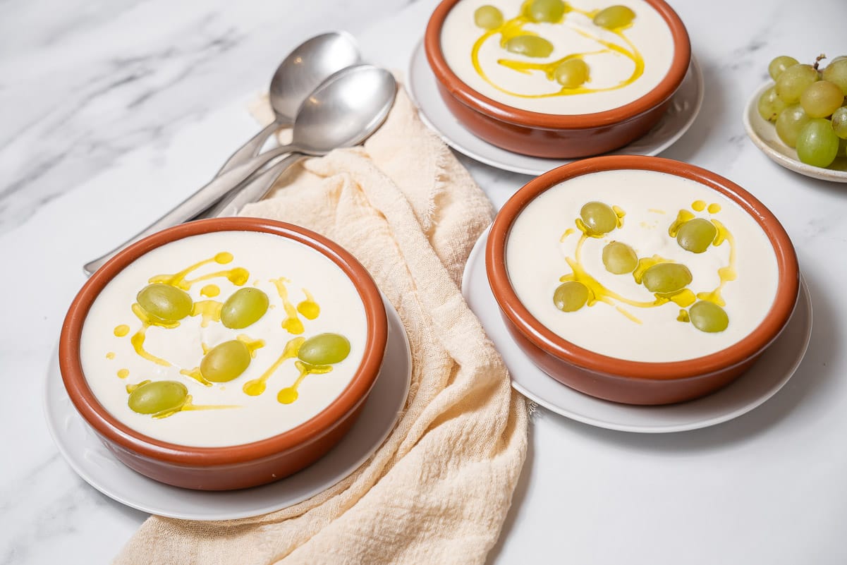 three clay dishes of ajoblanco topped with olive oil and grapes.
