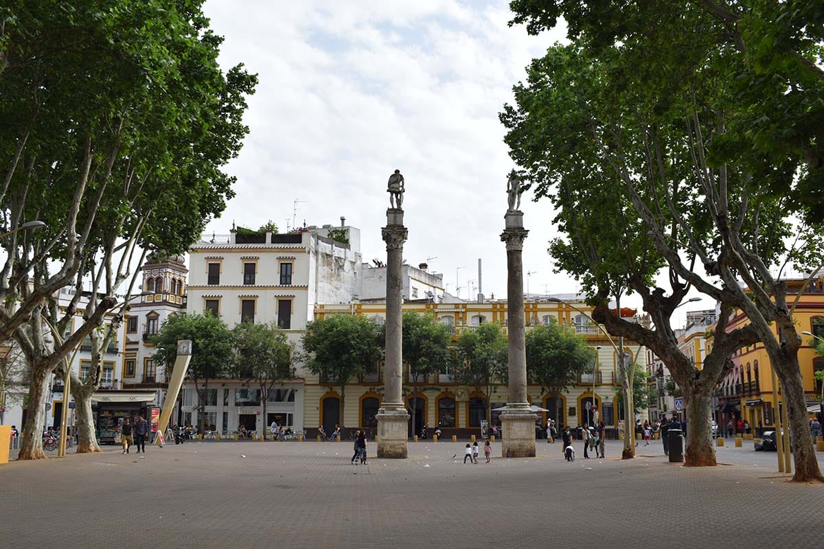 two columns in a tree-lined, paved square with buildings.