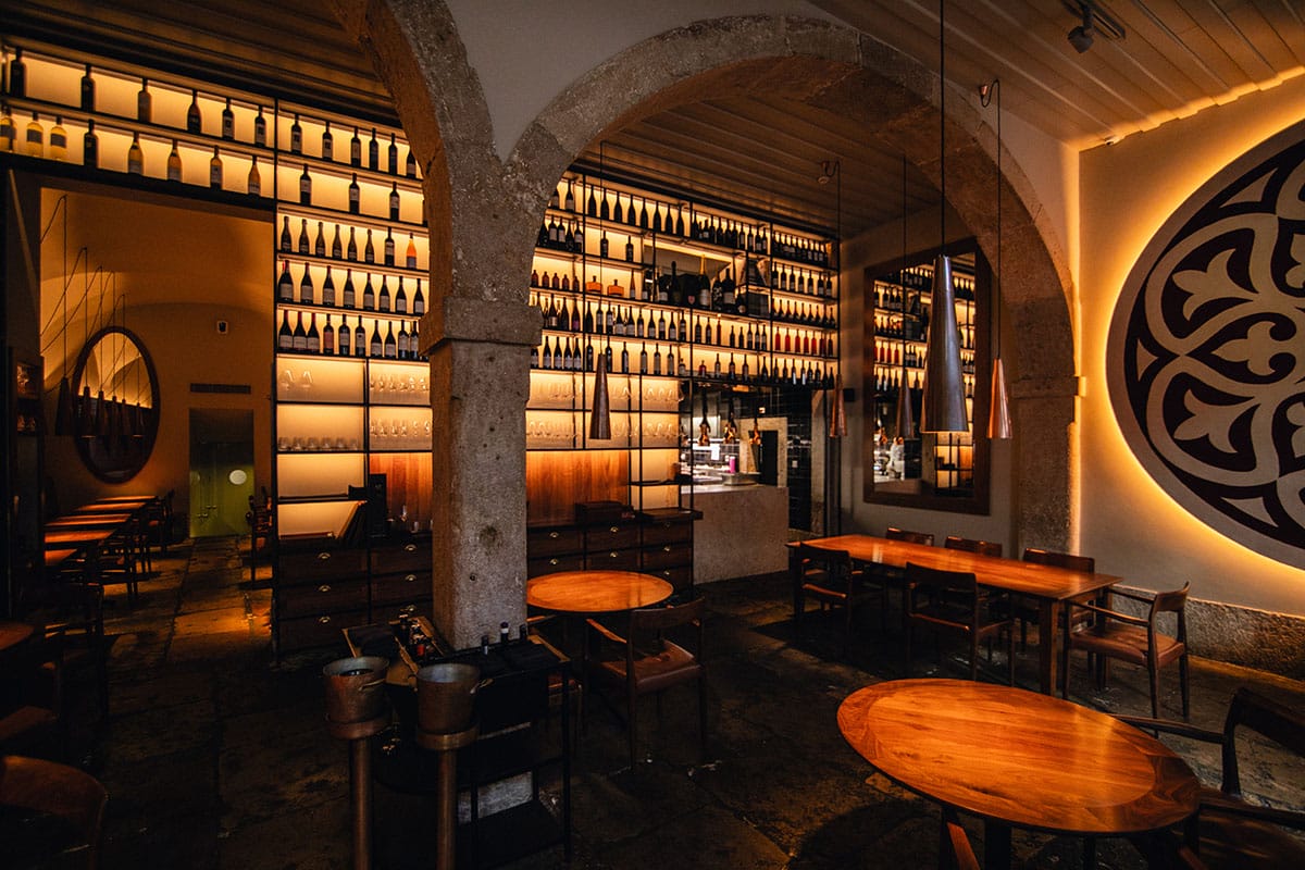 dark restaurant with round and rectangular tables and a wall lined with wine bottles on shelves.