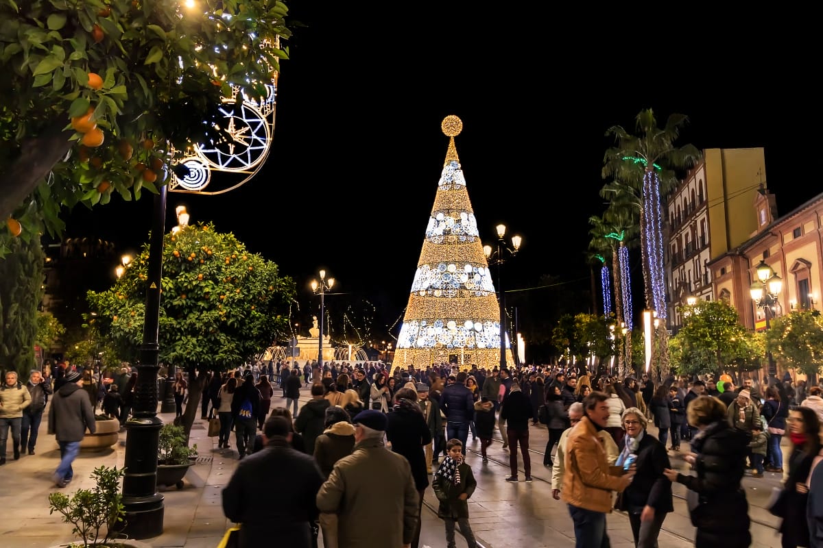 tall illuminated Christmas tree in a busy plaza surrounded by trees and buildings.
