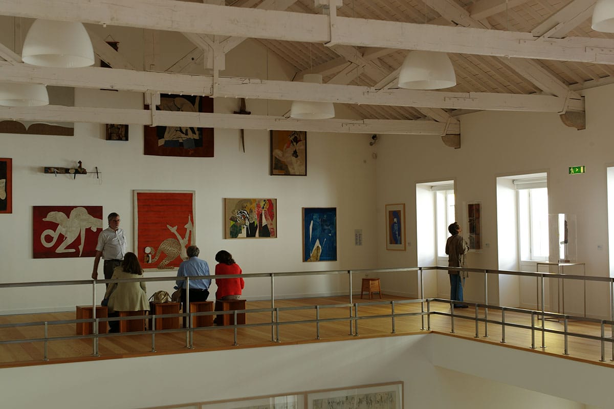 people sitting and standing as they look at paintings hanging on walls in an art museum.