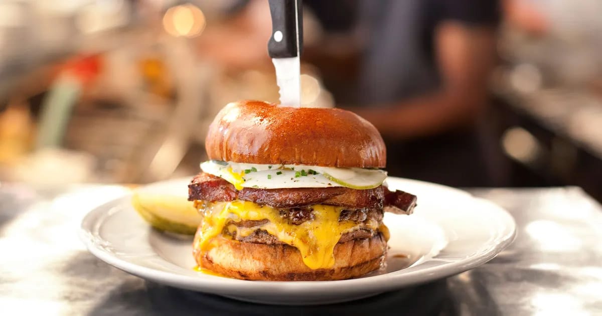tall cheeseburger with bacon and a fried egg with a steak knife inserted into it.