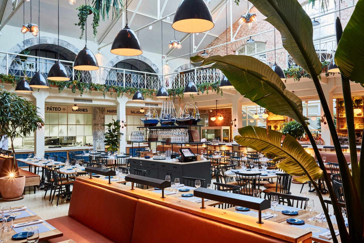 tables, chairs, and booths in a well-lit caf&eacute; with trees and model ships as decorations.