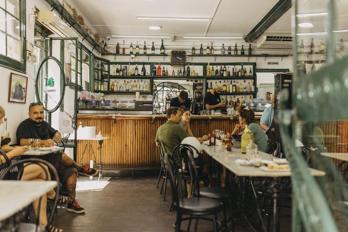 people sitting at tables in a small vermouth bar with shelves of bottles on the walls.