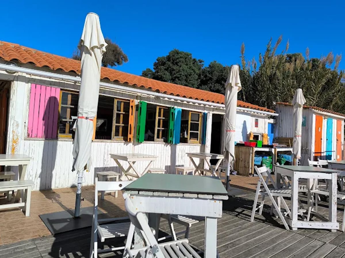 wooden tables and chairs outside of a white bar and restaurant with colorful shutters.