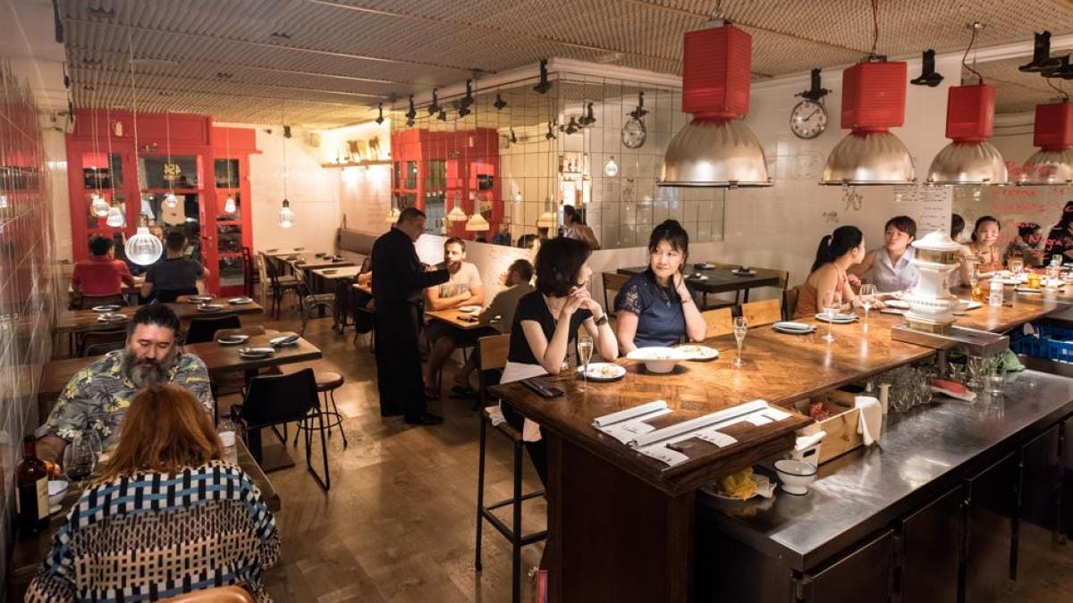 people sitting at a wooden bar and at small tables in a white restaurant with red accents on the chandeliers and walls.