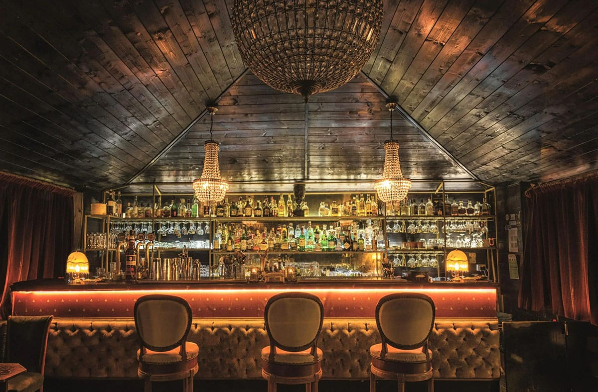 three bar stools at a bar with backlit shelves of liquor bottles under chandeliers.