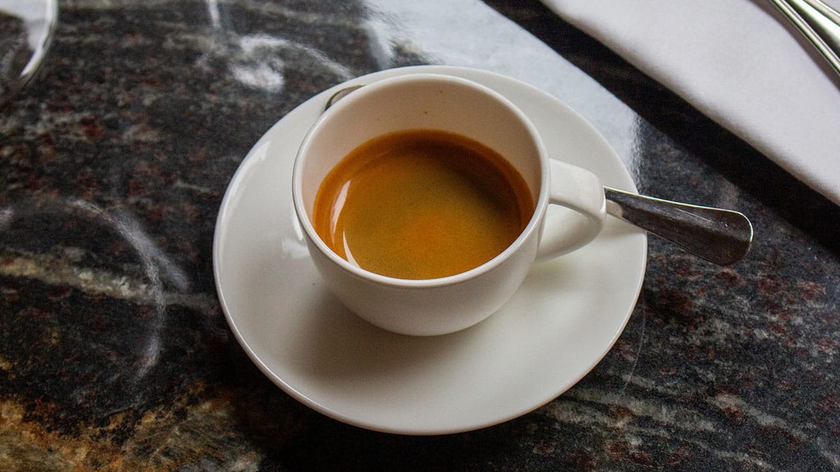 small white cup of espresso with a spoon and a napkin on a granite table.