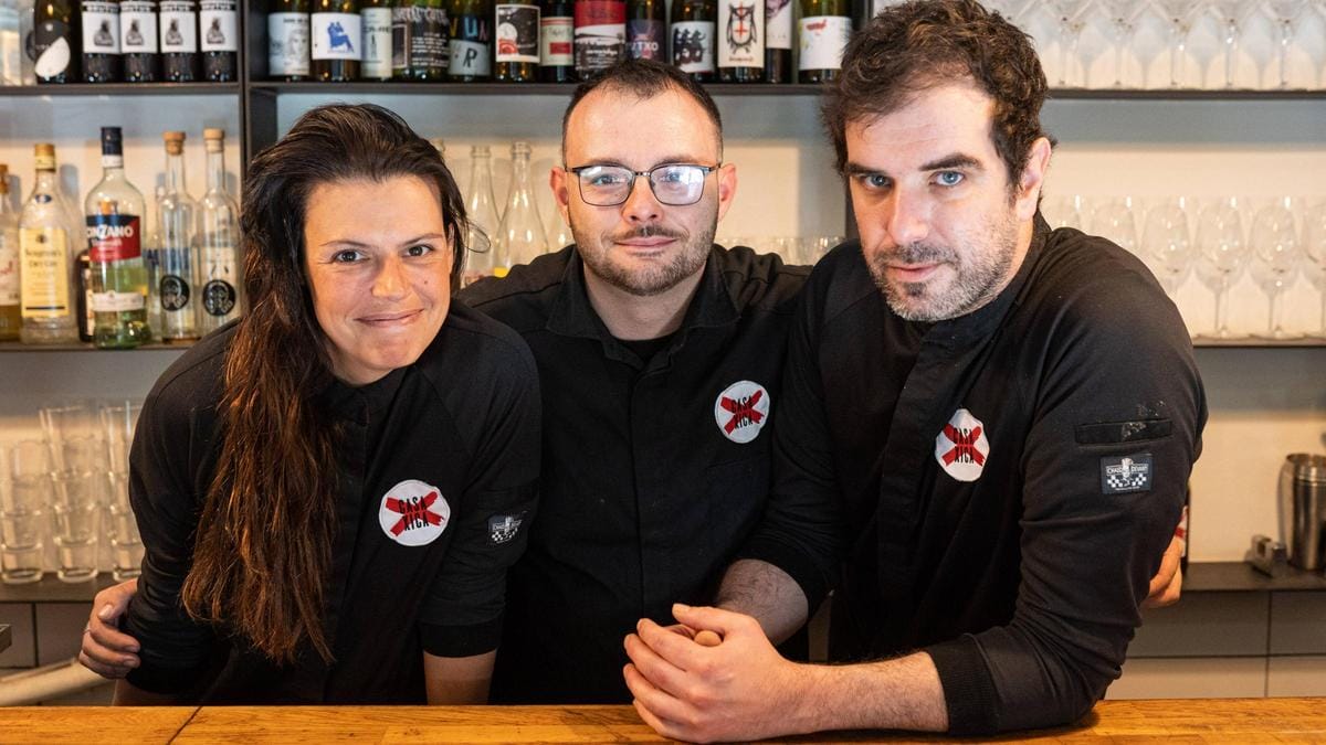 a woman and two men smiling behind a wooden bar with glasses and bottles behind them.