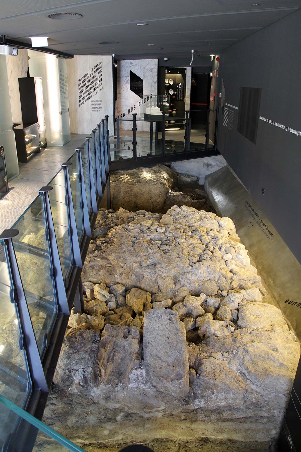 Roman stone ruins viewed from a glass-sided balcony.