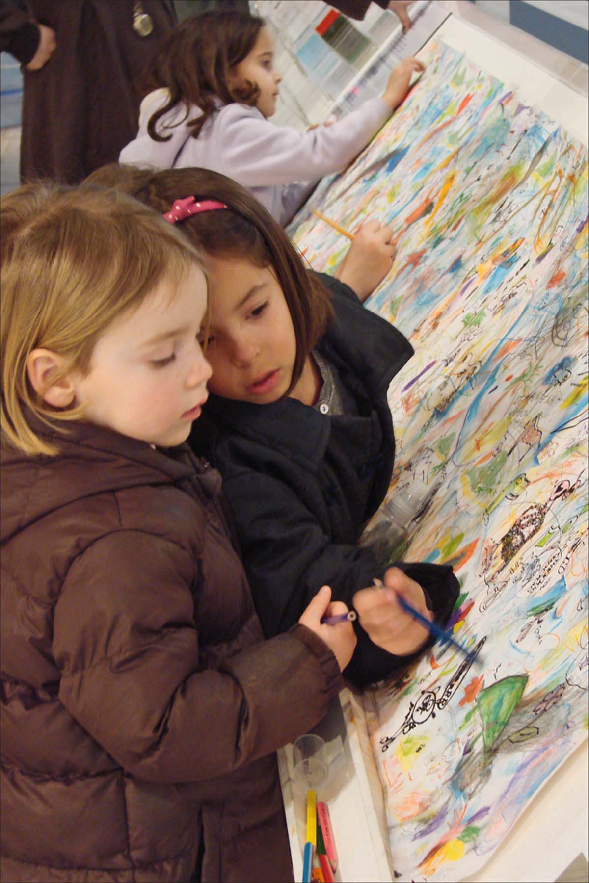 two girls drawing on a large white sheet of paper on a table.