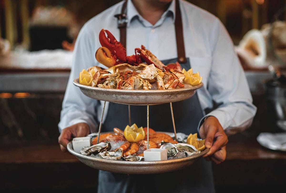 man wearing an apron and holding a two-tier tower of crabs, shrimp, and lobster.