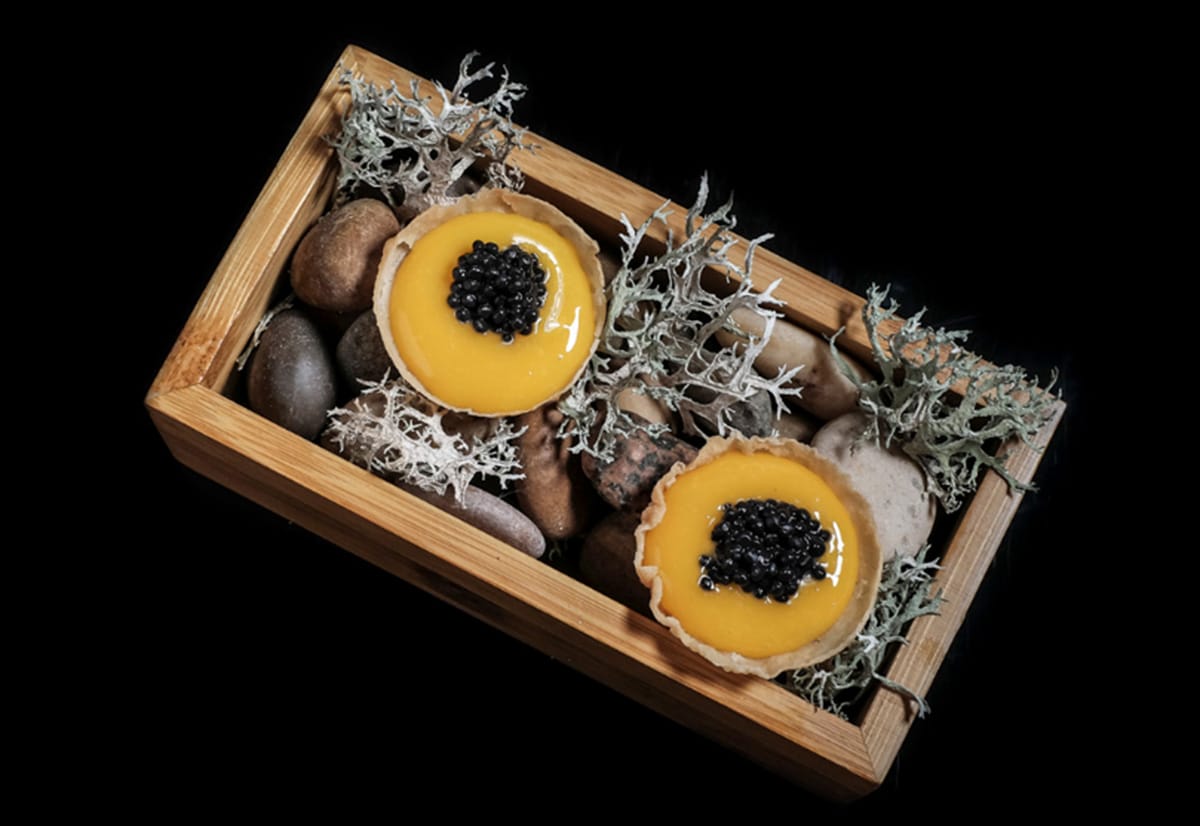 wooden box filled with rocks and seaweed and two dishes of yellow custard topped with caviar.