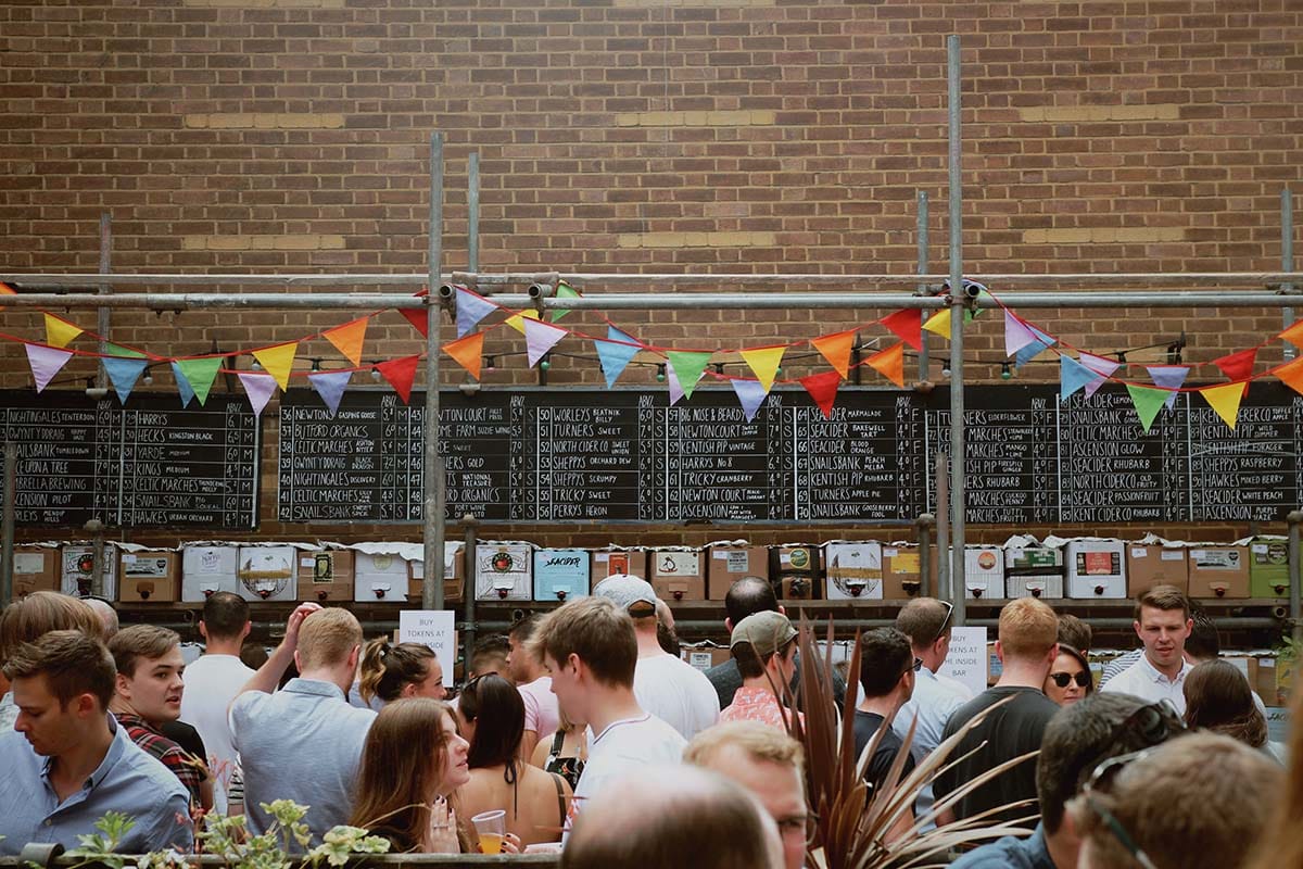 people at a festival with a brick wall behind them listing the types of ciders.