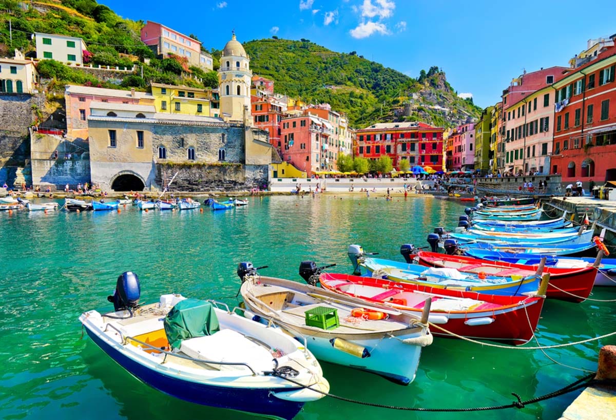 a row of colorful motor boats with a mountain and historic buildings in the background.