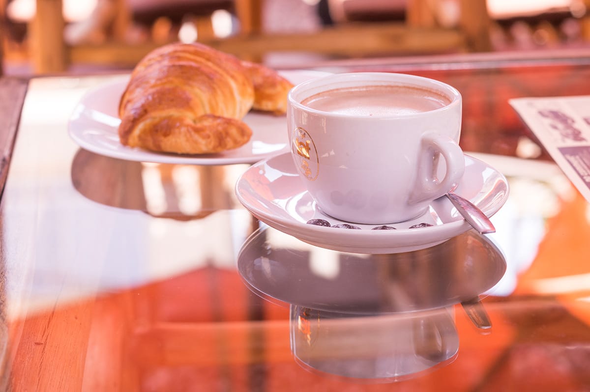 cup of coffee on a saucer with a spoon near a large croissant on a plate.