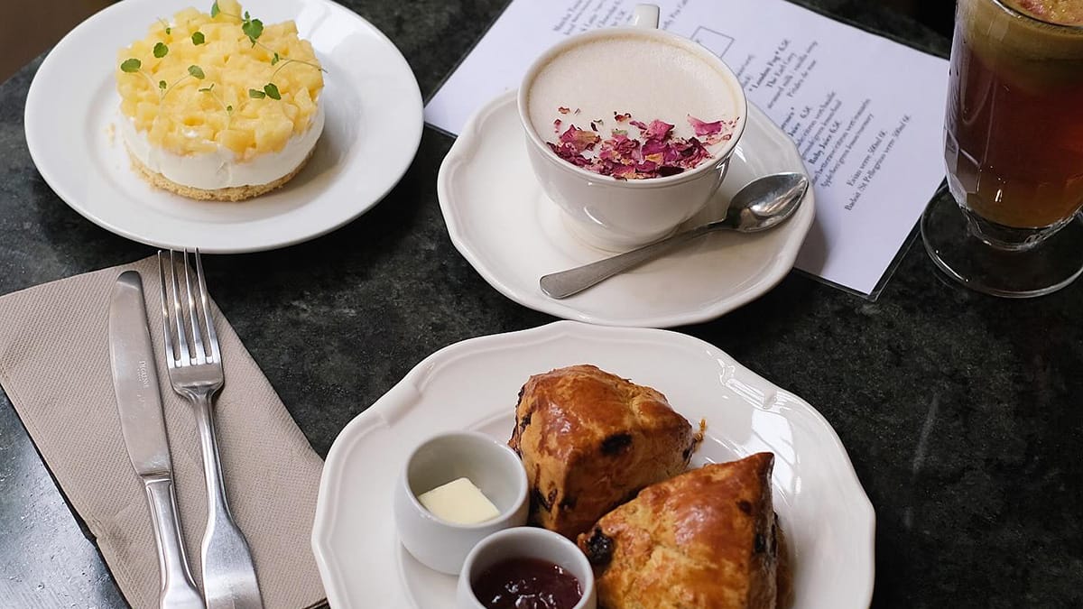 rose petal-topped latte with two plates of baked goods, utensils, napkin, and a menu on a granite table.