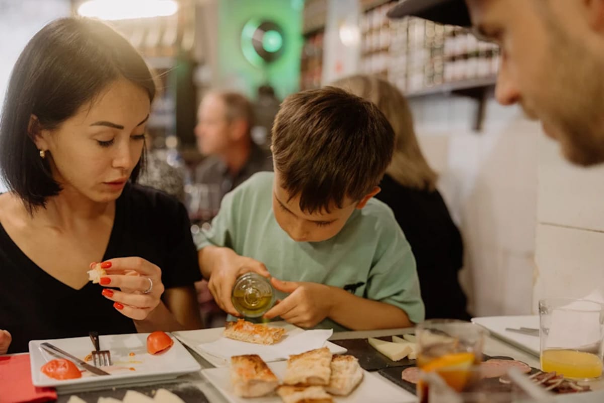 little boy pouring olive oil on toasted bread while his mom watches.