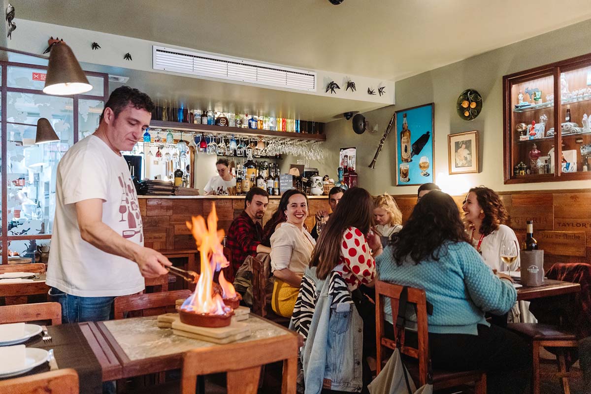 group of people sitting a table and watching flaming food in a clay pan.