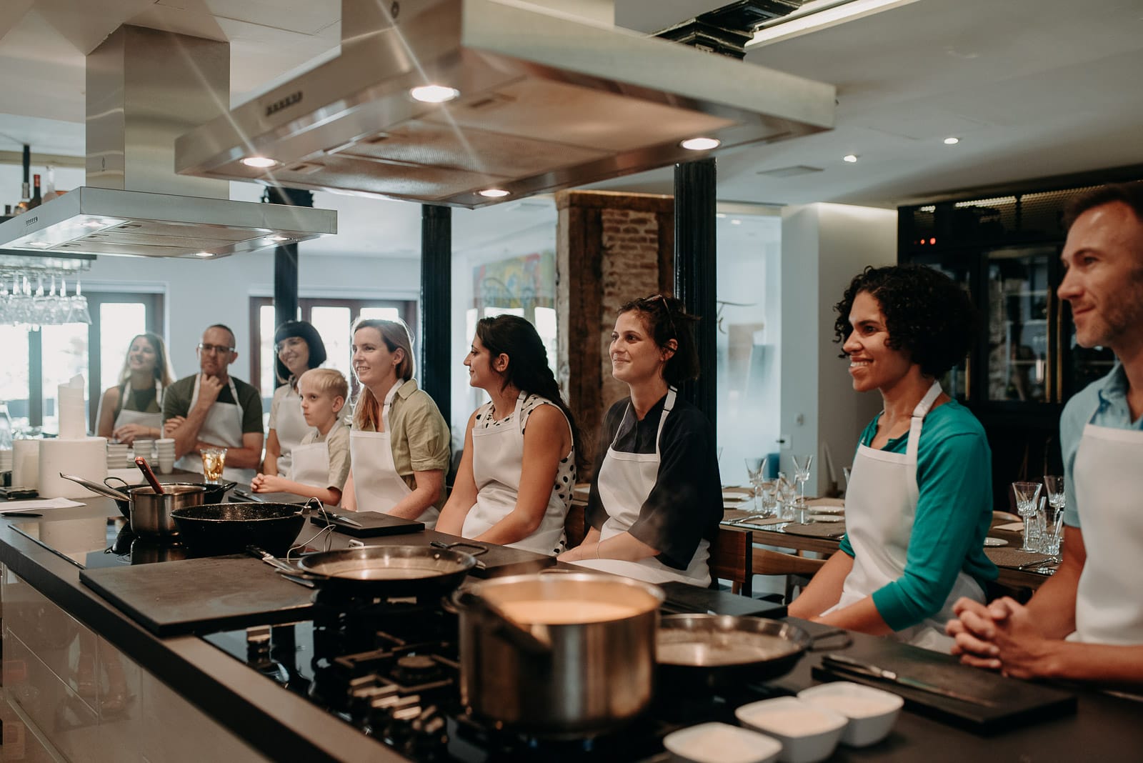 A group of people in aprons attentively watching a cooking demonstration in a modern kitchen with professional equipment.