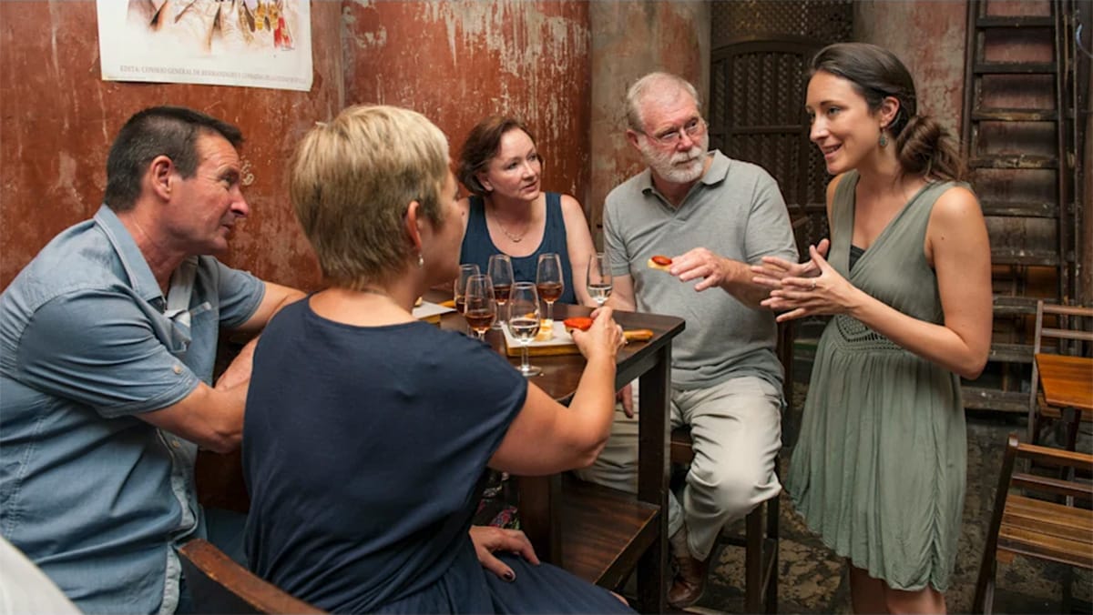people eating tapas and drinks at a table while a lady talks to them.