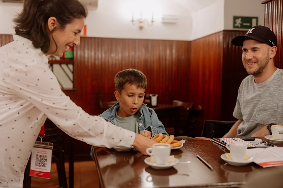 A smiling tour guide serves churros to a boy and his father at a café table with coffee cups.