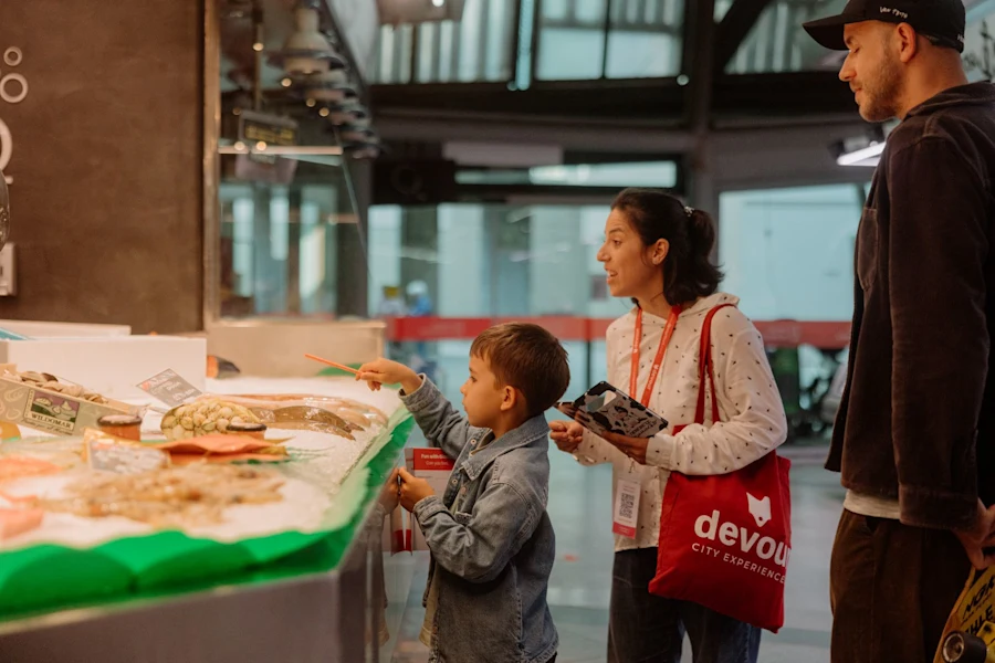 A young boy points at seafood in a market display while the guide explains and the father listens.