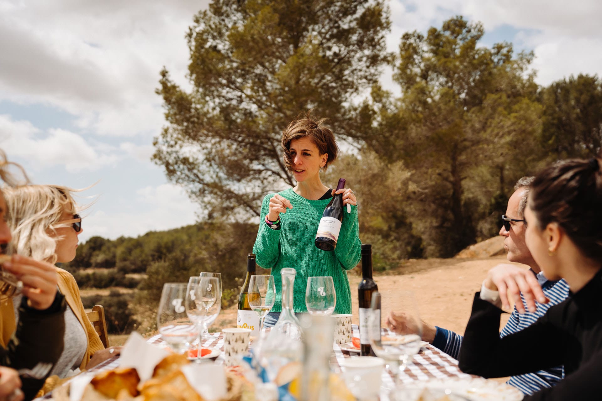 A woman holding a bottle of wine is enthusiastically explaining the features of the wine while standing near a table with glasses and bottles. The picturesque vineyard backdrop adds to the warmth of the moment.