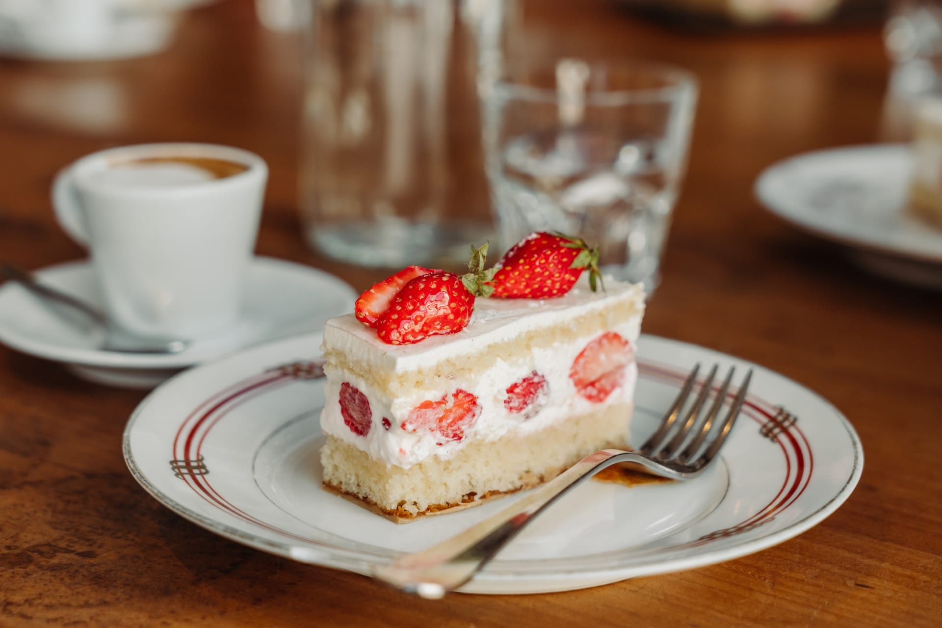 A slice of strawberry shortcake with whipped cream and fresh strawberries on top, served on a white plate with red detailing, next to a cup of coffee and a glass of water.
