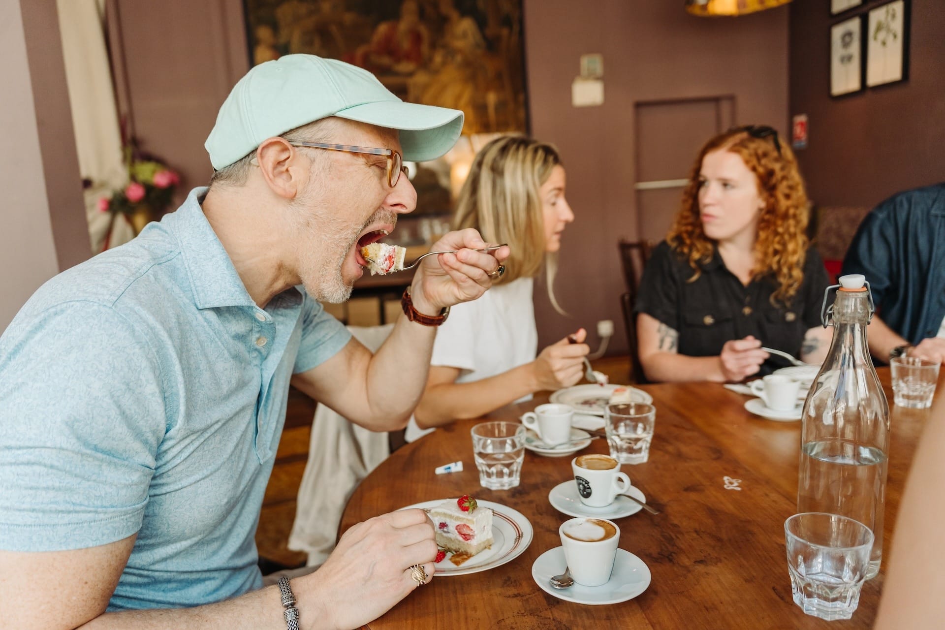 A man in a mint-green cap takes a big bite of strawberry cake while sitting at a wooden café table with espresso cups and friends chatting in the background.