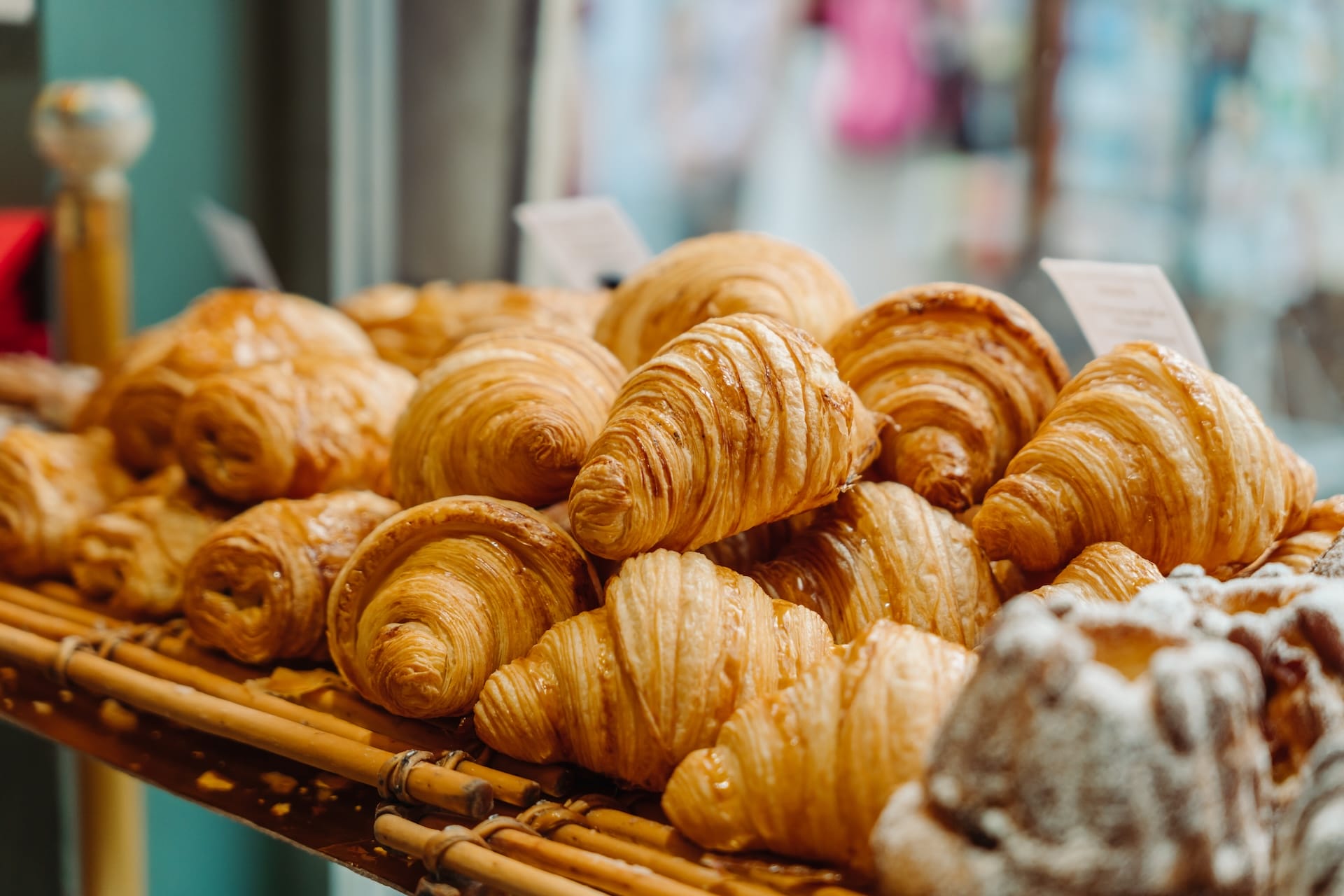 Close-up of a bakery display filled with golden, flaky croissants and pastries, arranged on a wooden rack behind a glass window.