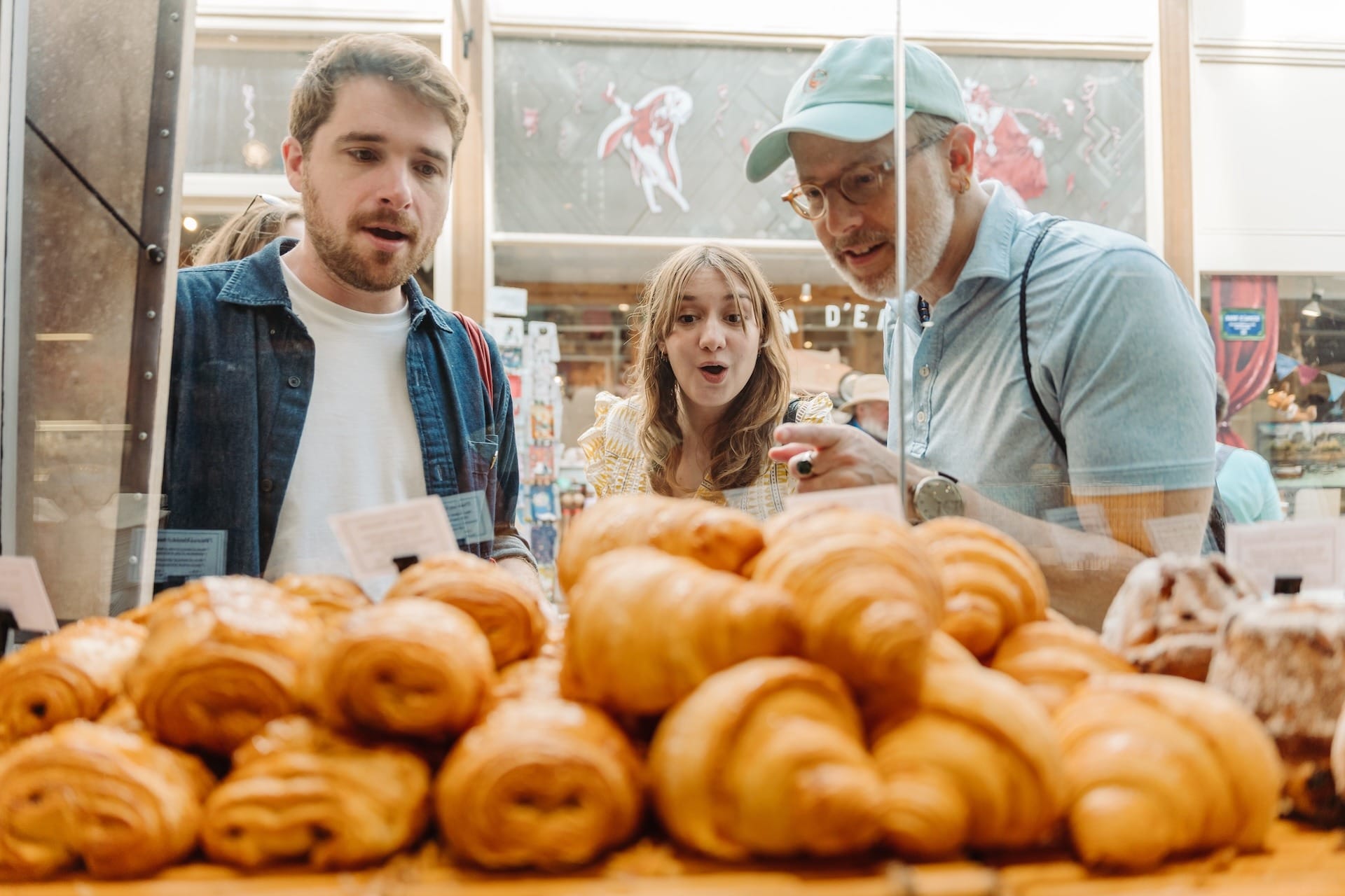 Three people looking with excitement through a bakery display case filled with golden croissants and pastries.