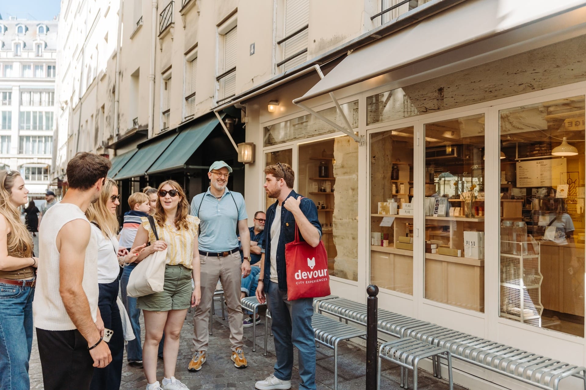 A group of people standing outside a café or shop on a narrow city street, listening to a guide holding a red “Devour City Experiences” tote bag.