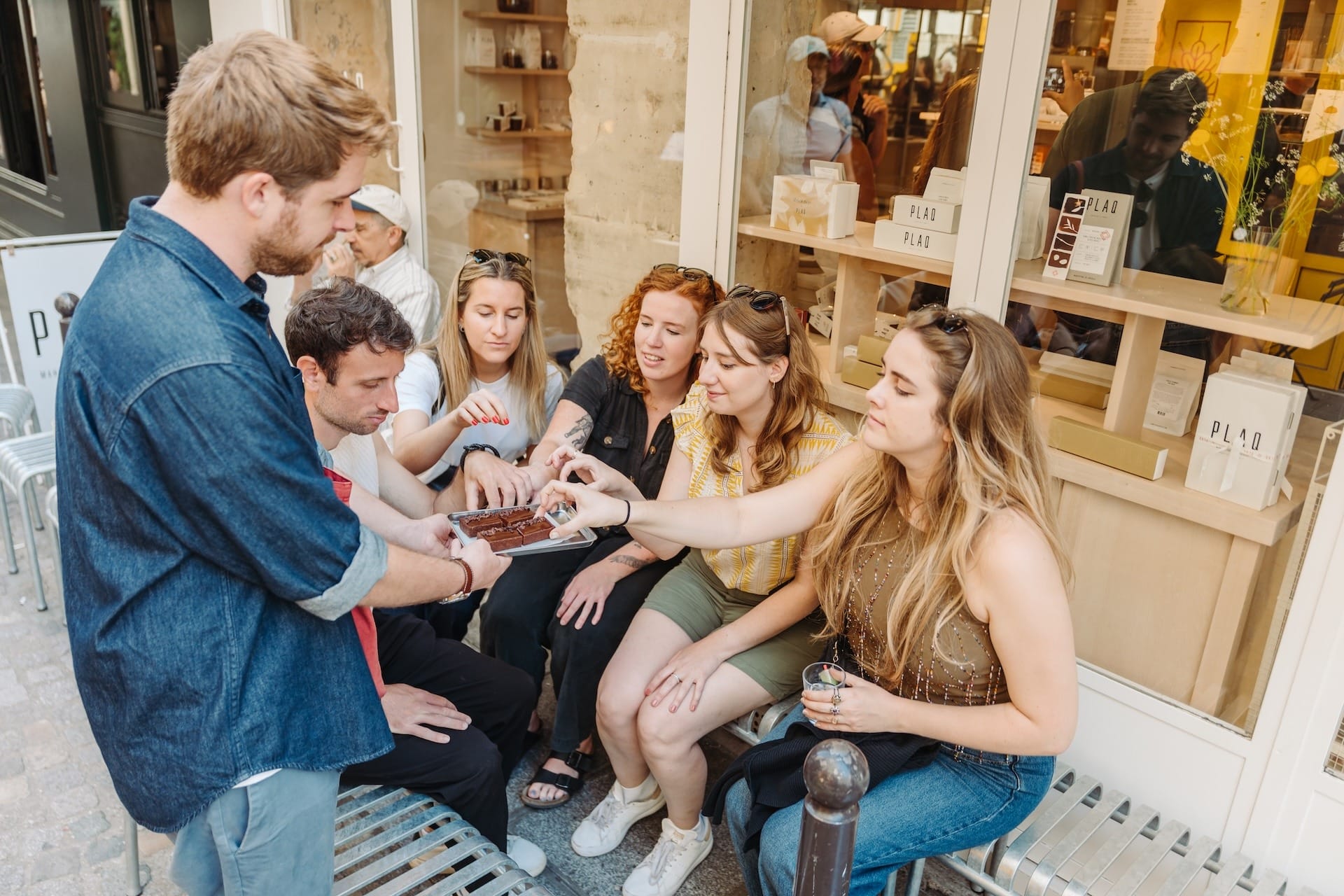 A group of people sitting outside a shop, eagerly reaching for pieces of chocolate being offered on a tray by a guide.