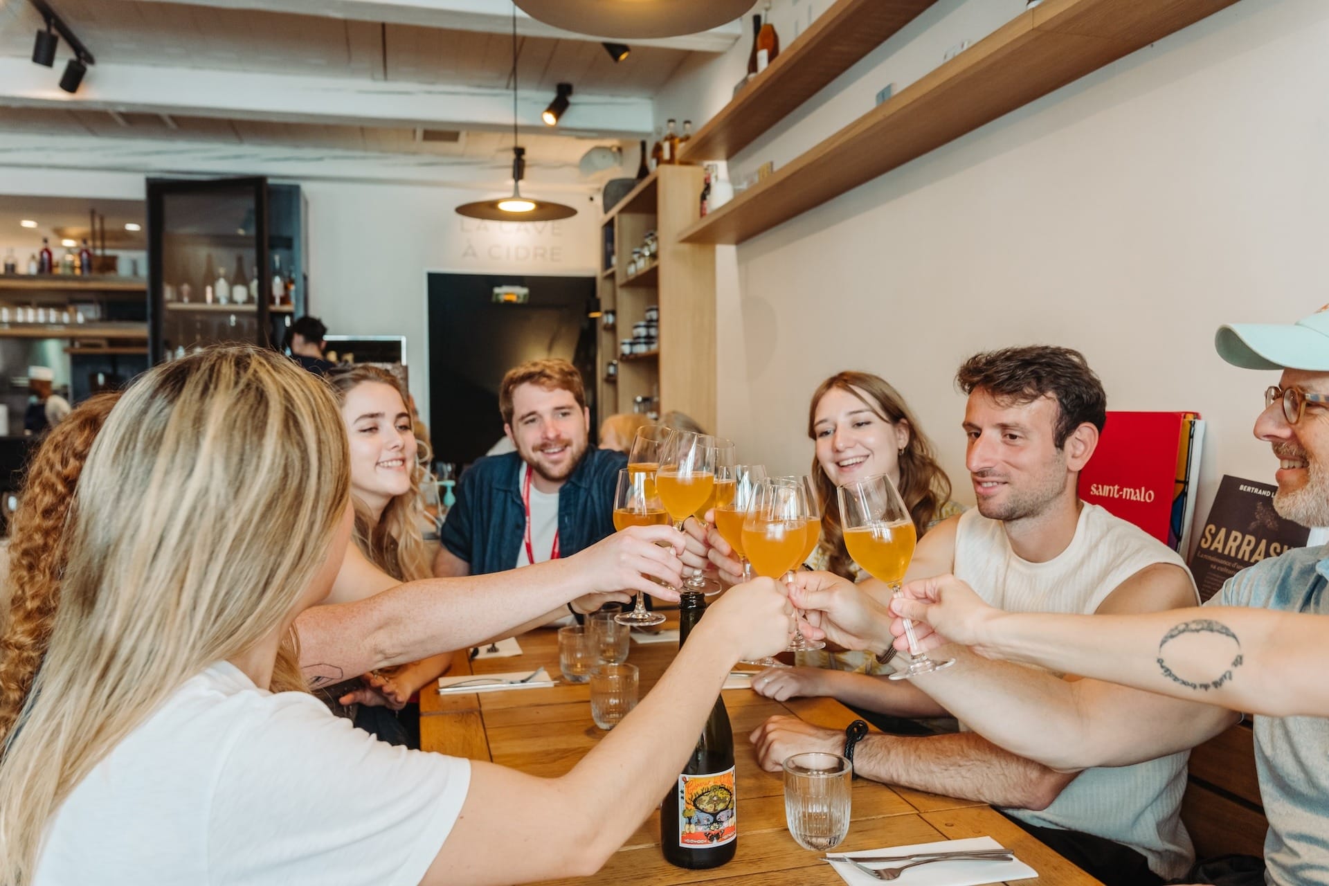 A group of friends sitting around a wooden table in a restaurant, clinking glasses of golden cider together with big smiles.