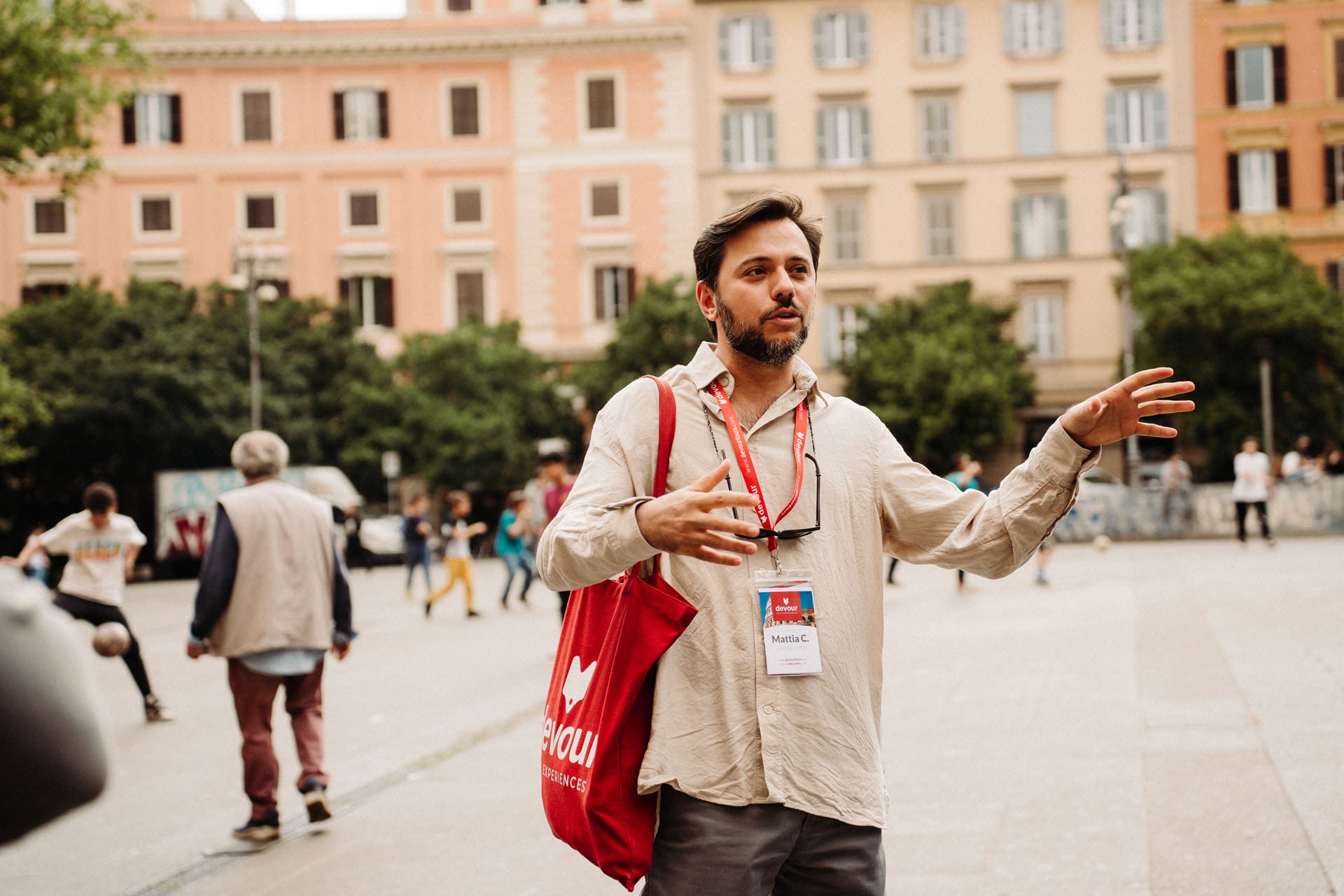 A tour guide with a red Devour tote bag speaks animatedly to a group in a lively Italian square.