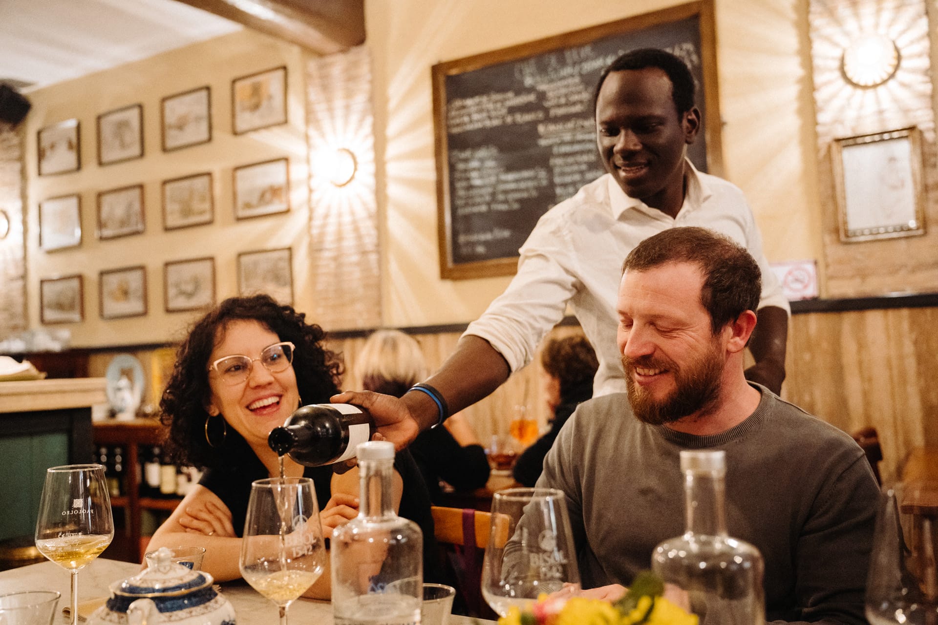 A server pours wine into a guest’s glass at a cozy Italian restaurant, while two diners smile warmly.