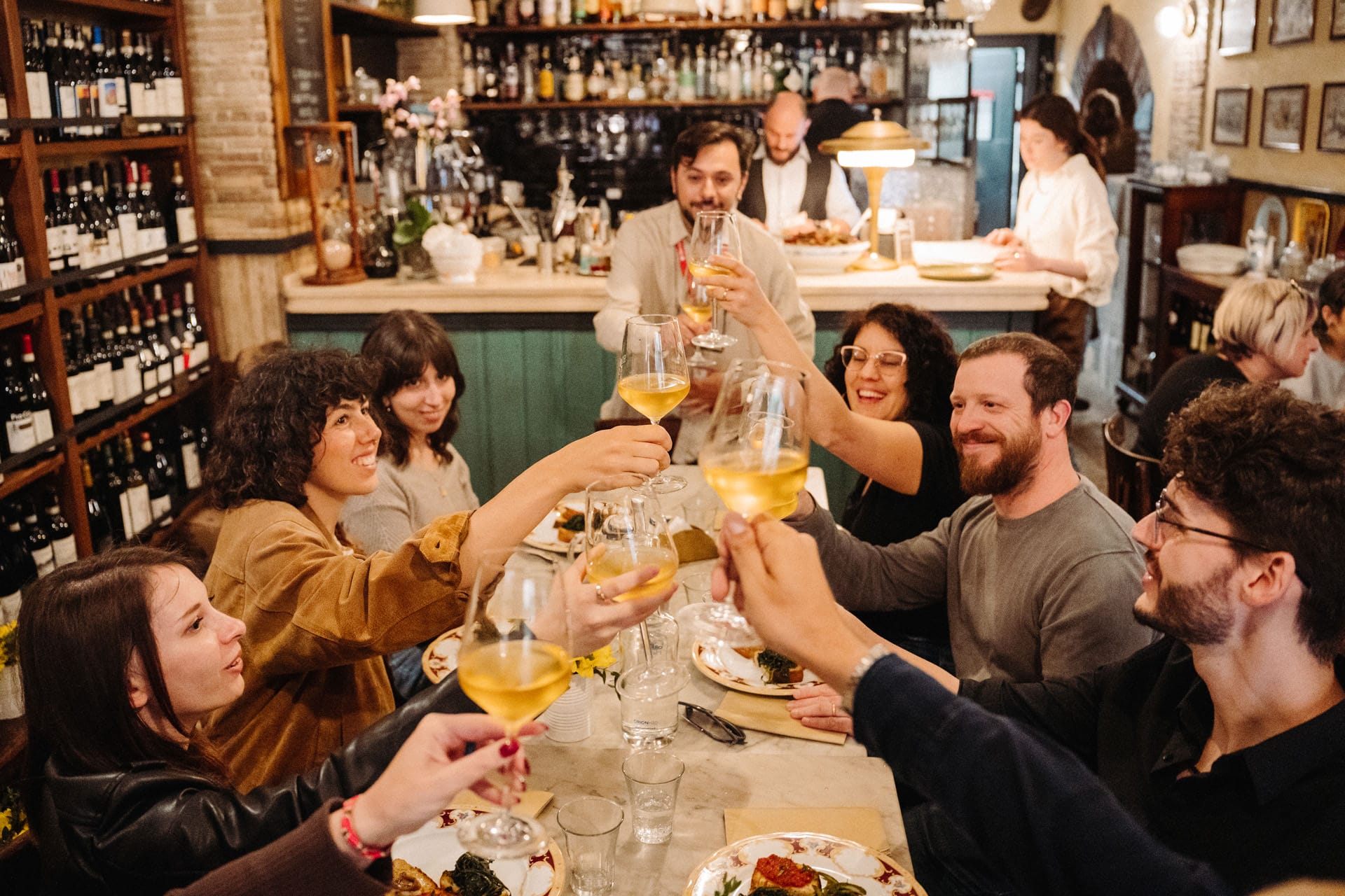 A group of diners raises glasses of white wine in a toast inside a rustic wine bar.