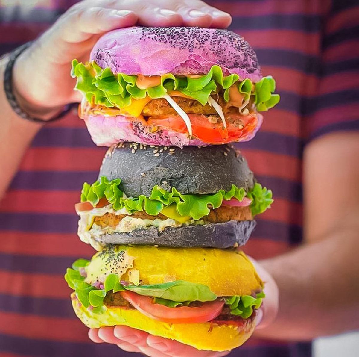person holding a stack of three colorful vegan burgers.