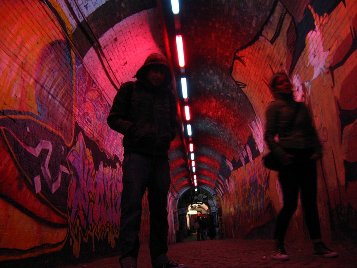 people walking down a brick tunnel covered with graffiti and illuminated with red and white lights.