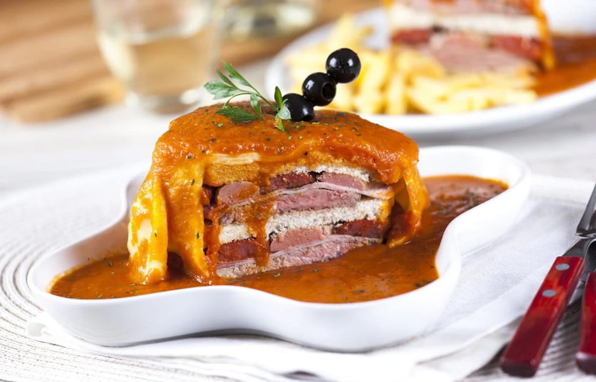 tall sandwich layered with bread, meat, and cheese in a large white dish topped with tomato sauce.