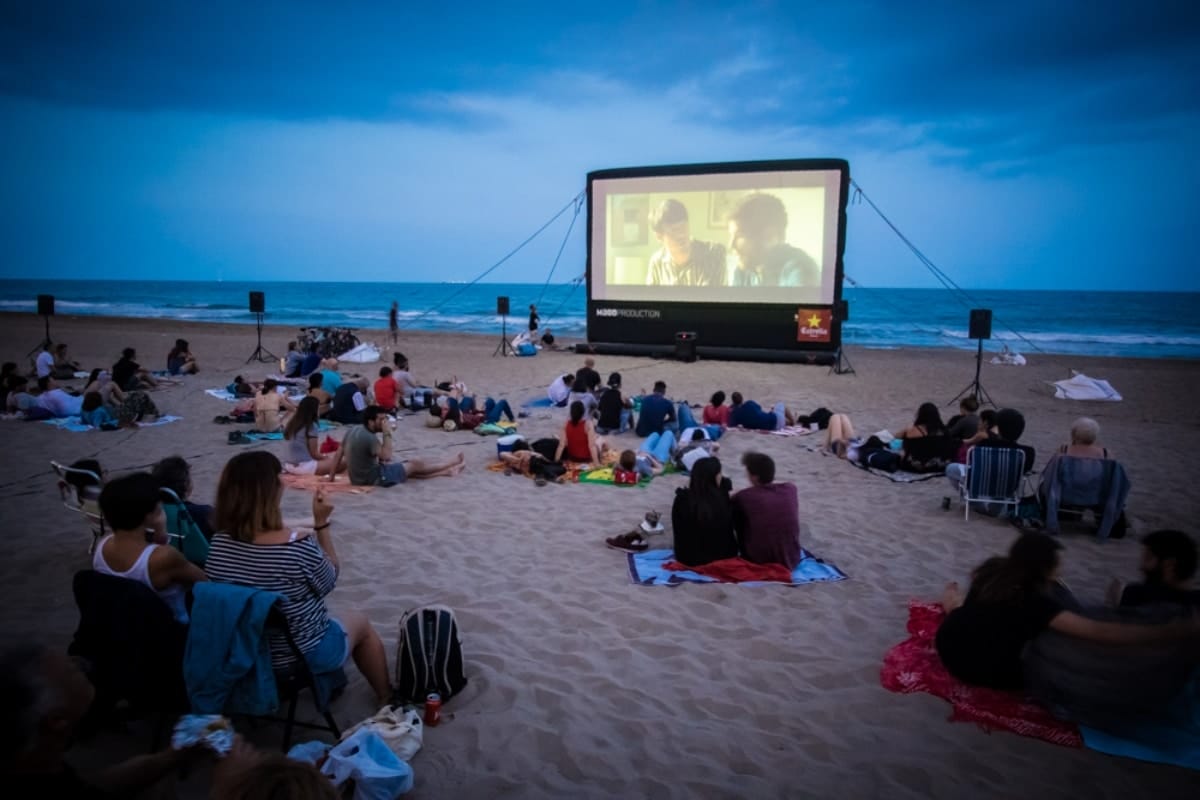 people sitting on chairs and towels on the beach while watching a movie projected onto a large screen with the ocean in the background.