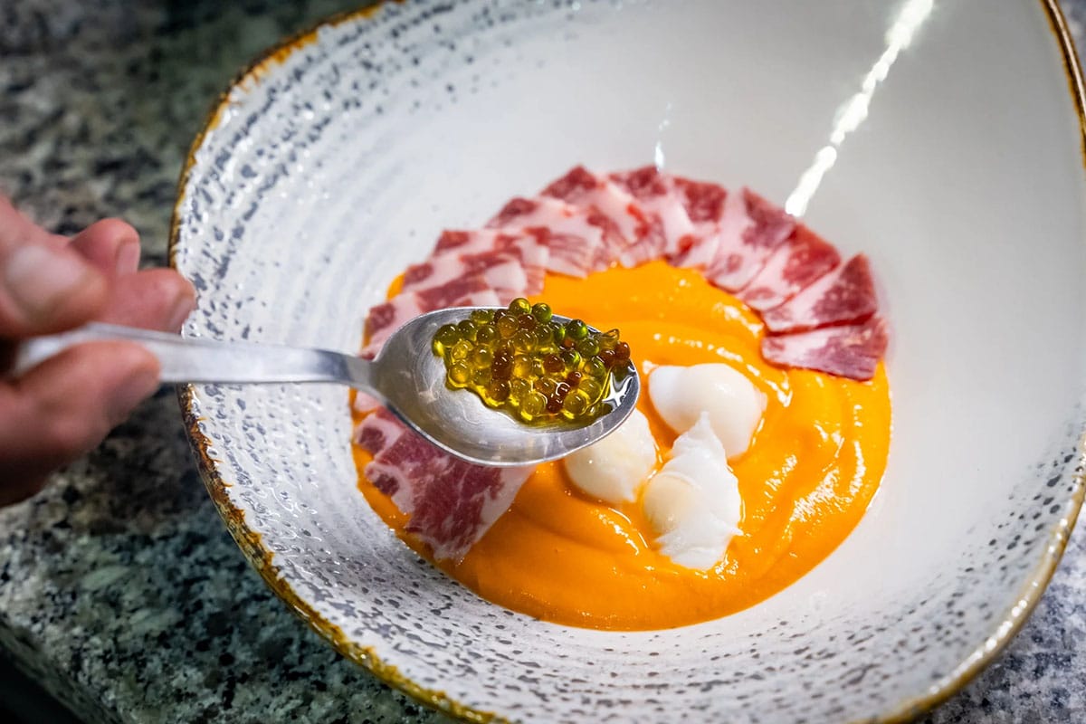 holding a spoonful of caviar with a bowl of salmorejo garnished with ham.