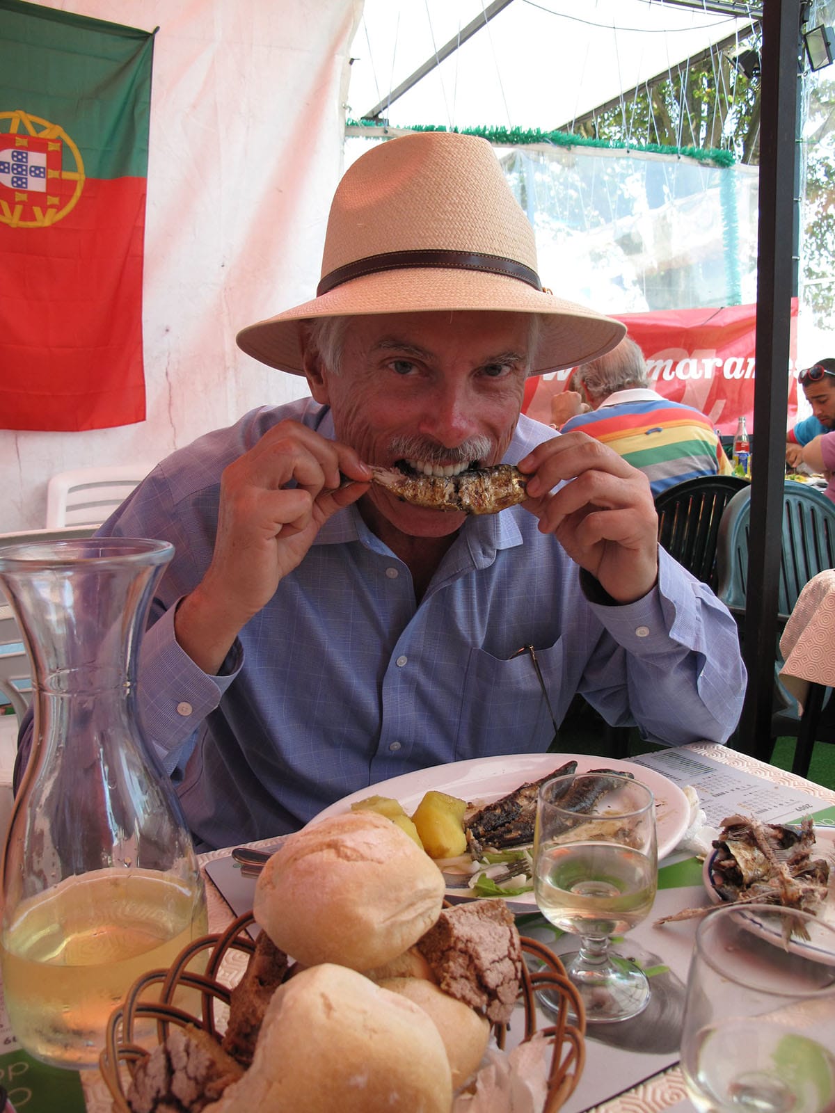 man wearing a hat and eating grilled sardines outdoors at a table.