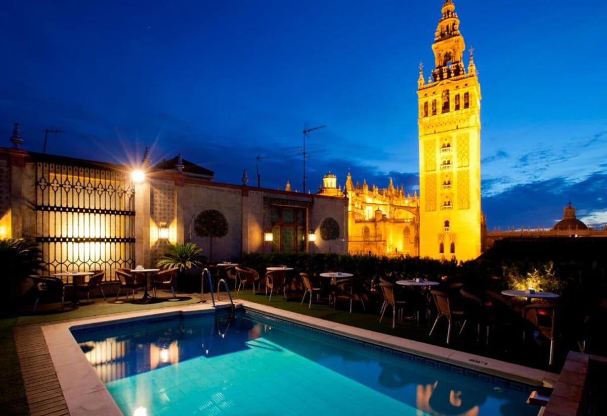 rooftop pool and terrace overlooking an illuminated cathedral tower at sunset.