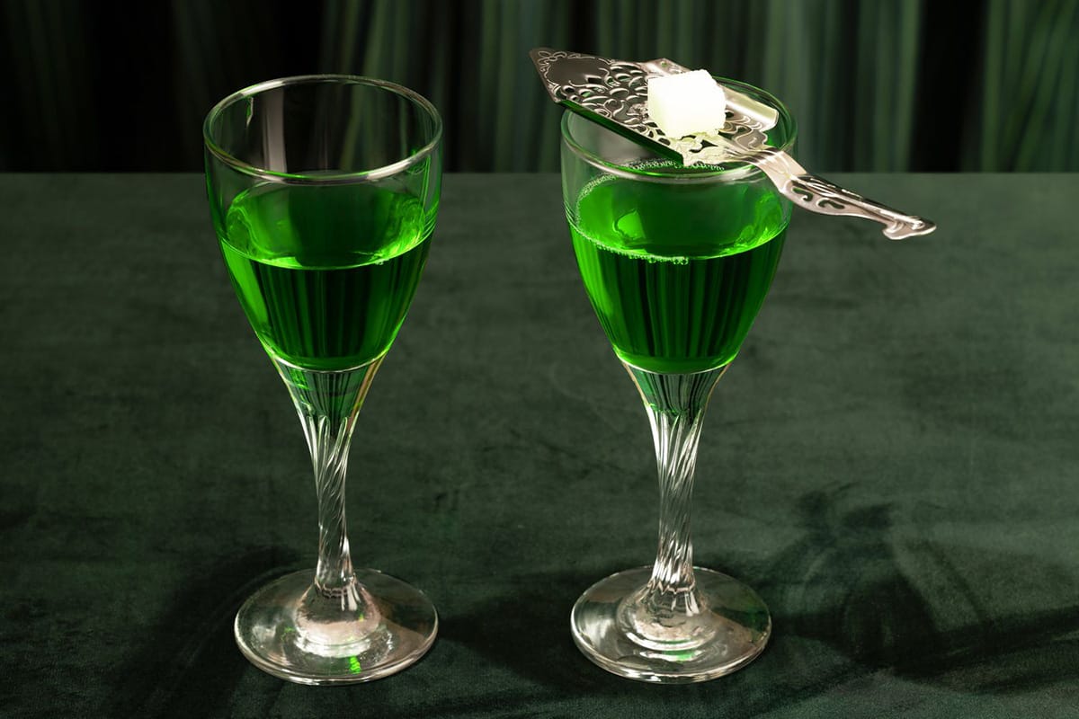 two stemmed glasses of abisinthe with a sugar cube on a silver utensil.