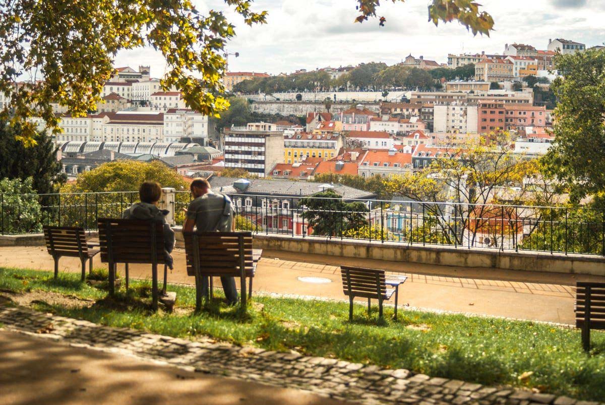 two people sitting on wooden park benches in a green space overlooking Lisbon.